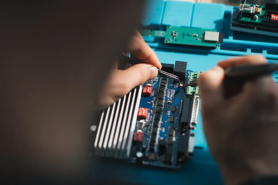 Hands using tweezers and a soldering iron to work on a circuit board on a blue electronics mat.